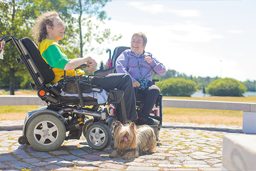 Kaksi hymyilevää pyörätuolin käyttäjää koiran kanssa kesällä ulkona.