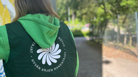Pitkähiuksinen hainen selkä päin kameraa, hupparin selässä lukee Hyvän arjen rakentajat